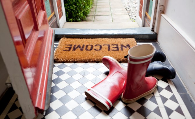 A welcome door mat by an open door next to a pair of red wellies